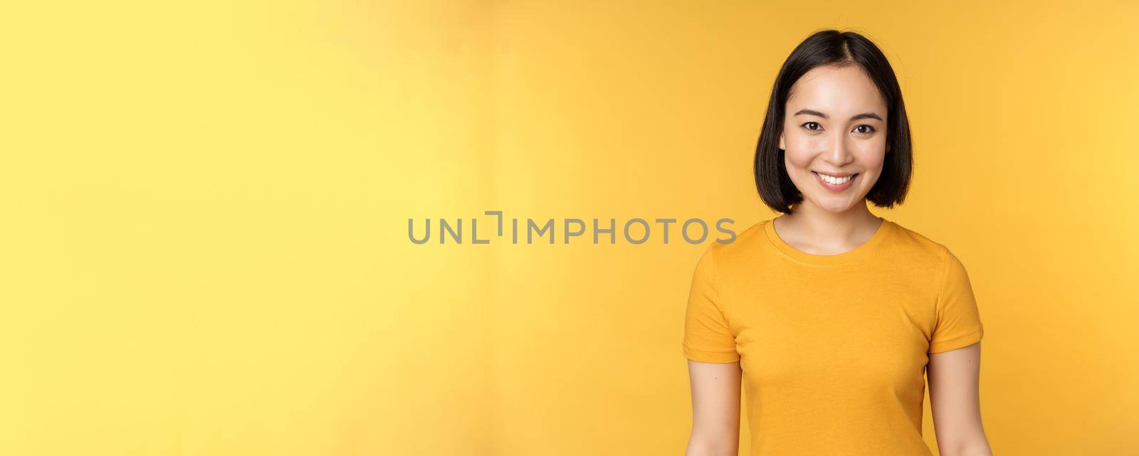 Portrait of young modern asian woman, smiling happy with white teeth, looking confident at camera, wearing casual t-shirt, standing over yellow background by Benzoix