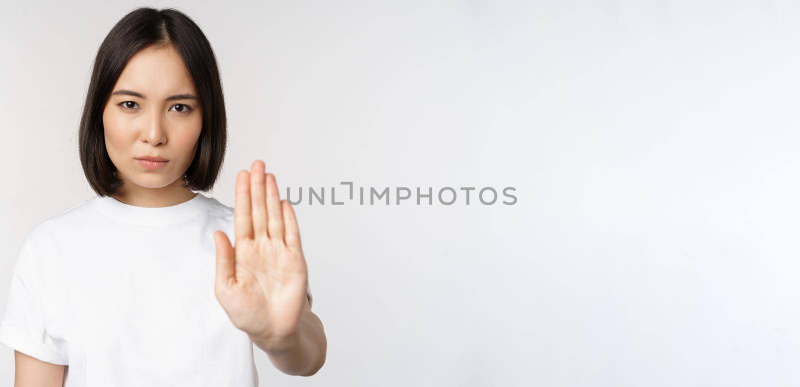 Image of asian girl showing stop, prohibit smth, extend one arm to show forbidding, taboo gesture, standing in tshirt over white background by Benzoix