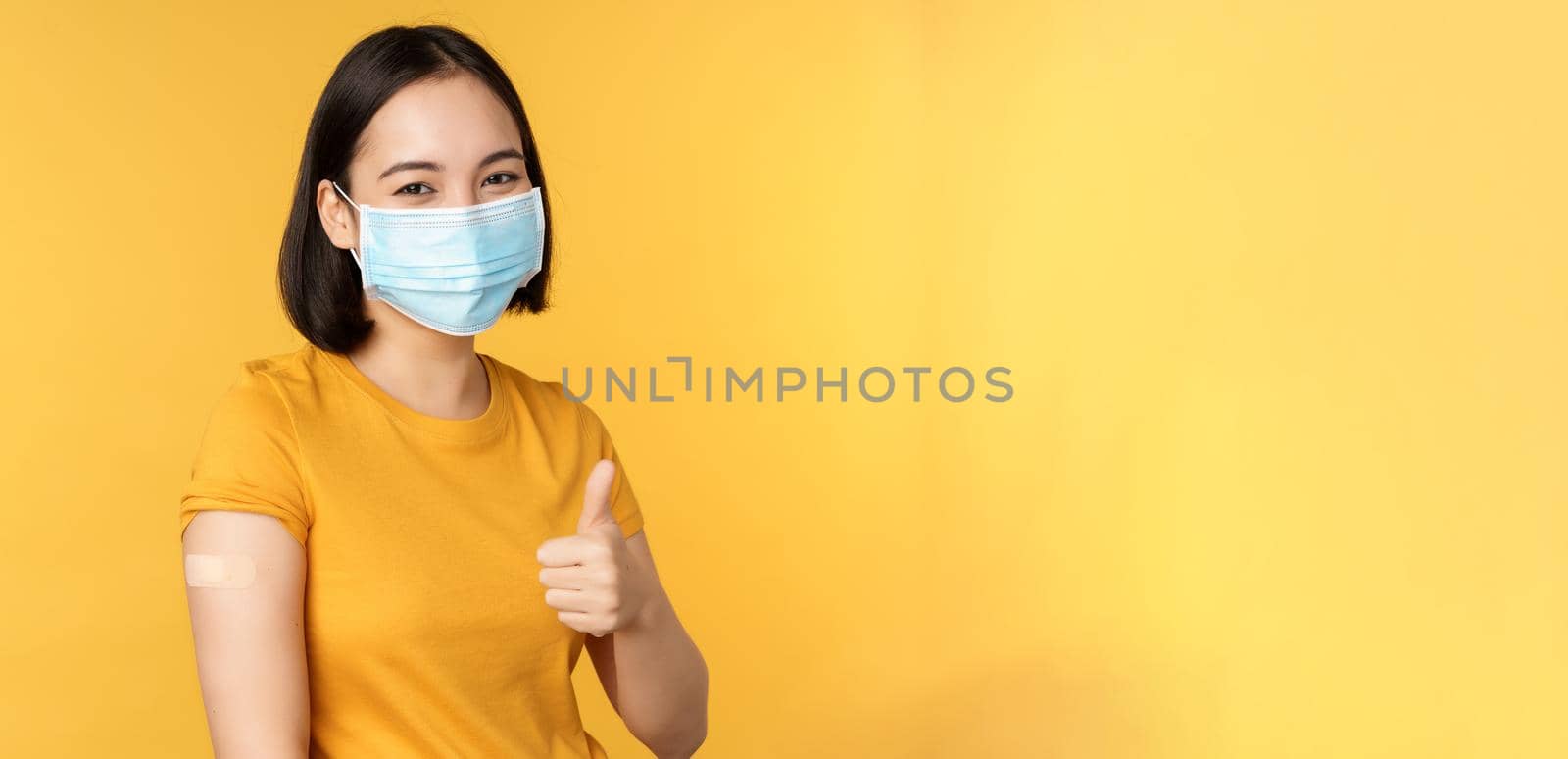 Vaccination from covid and health concept. Happy asian girl showing thumbs up, wearing medical mask, band aid on shoulder, got coronavirus vaccine shot, yellow background.