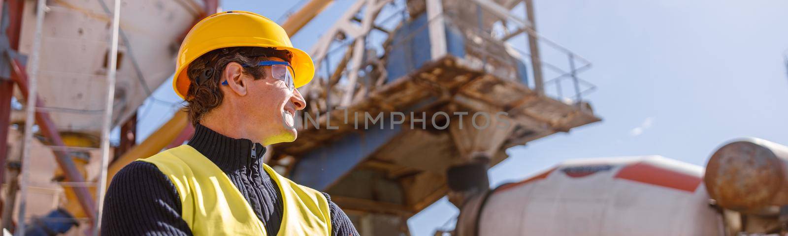 Cheerful matured man wearing work vest and safety helmet while keeping arms crossed and looking at factory machine