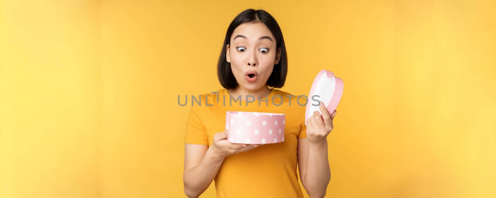 Portrait of excited asian woman, open gift box with surprised happy face, standing over yellow background.