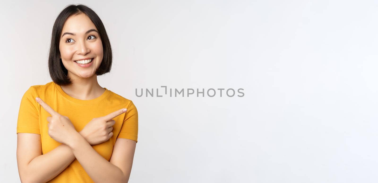 Cute asian girl pointing fingers sideways, showing left and right promo, two choices, variants of products, standing in yellow tshirt over white background.