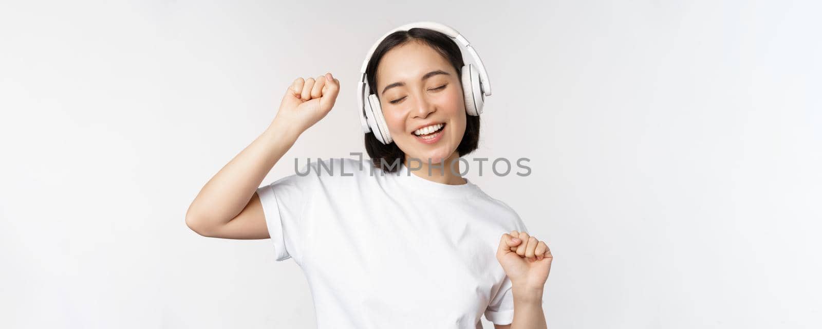 Dancing and singing asian woman, listening music in headphones, standing in earphones against white background by Benzoix