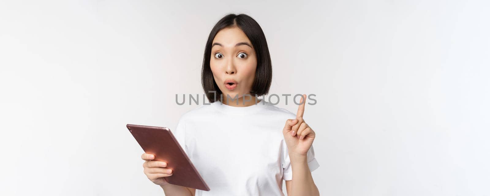 Enthusiastic asian woman with tablet, raising finger and looking amazed, pointing up, standing over white background.