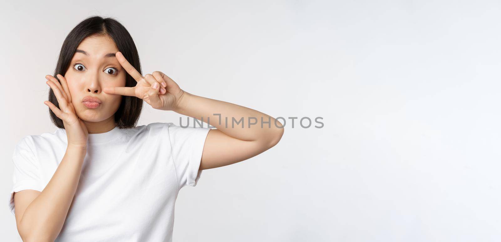 Cute asian girl posing with kawaii v-sign, peace gesture near face, standing in tshirt over white background. Copy space