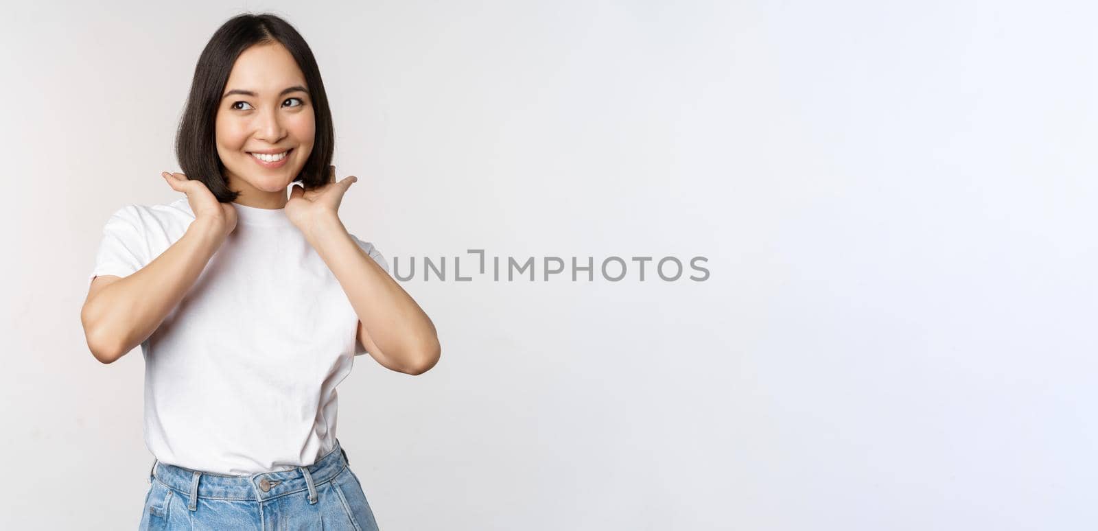 Portrait of cute, beautiful asian woman touching her new short haircut, showing hairstyle, smiling happy at camera, standing over white background.