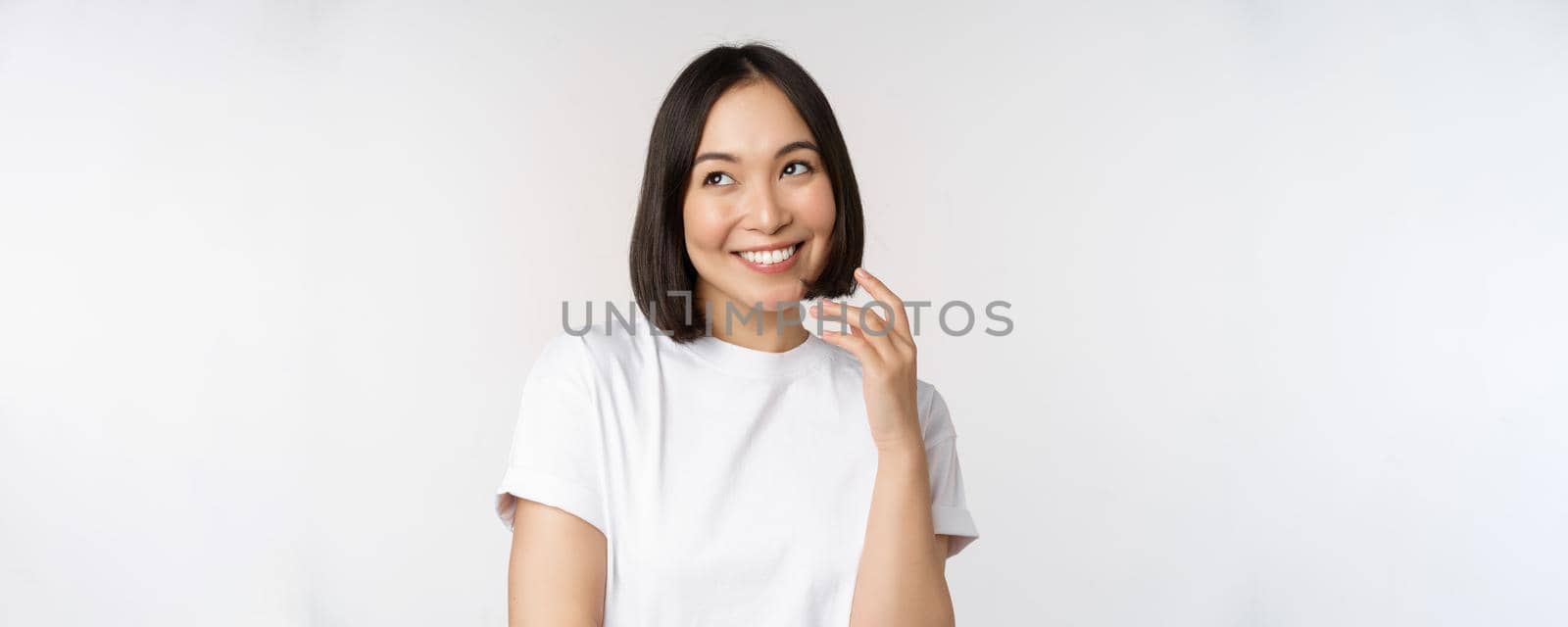 Portrait of cute coquettish woman laughing and smiling, looking aside thoughtful, thinking or imaging smth, standing in white t-shirt over studio background by Benzoix