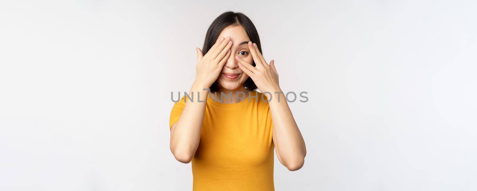 Cute korean female model, cover eyes with hands, peeking through fingers curious, watching smth, standing in yellow tshirt over white background.