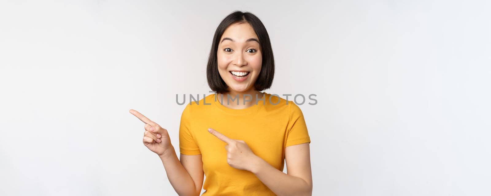 Portrait of smiling asian brunette girl in yellow tshirt, pointing fingers left, showing copy space, promo deal, demonstrating banner, standing over white background.