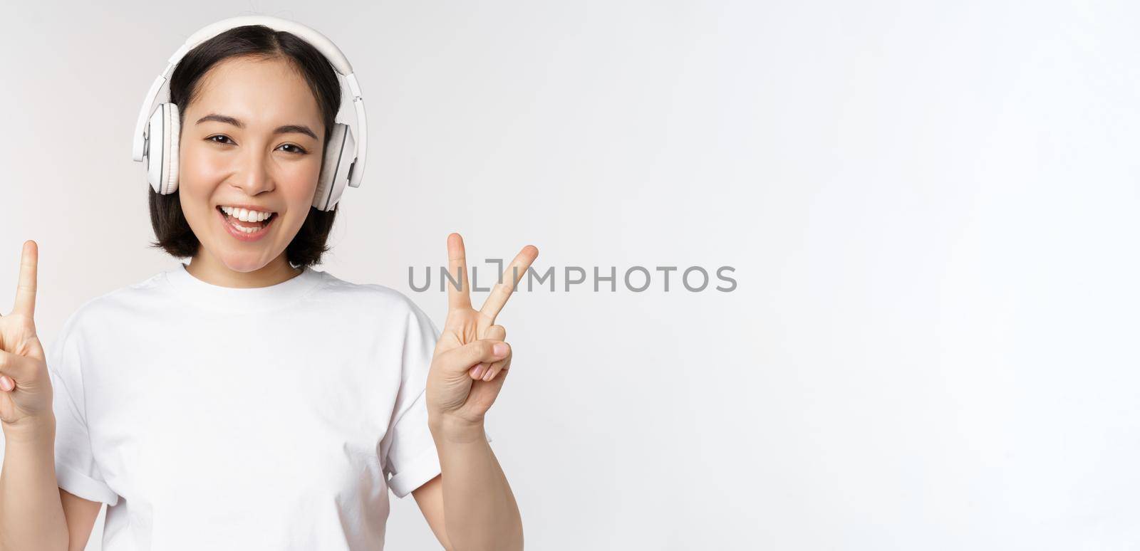 Happy asian woman wearing headphones and smiling, showing peace v-sign, listening music, standing in tshirt over white background by Benzoix