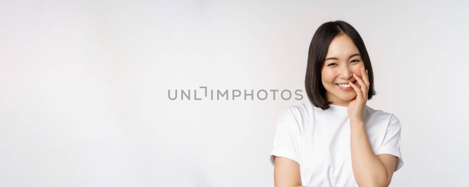 Portrait of young beautiful woman, korean girl laughing and smiling, looking coquettish, standing against white background.