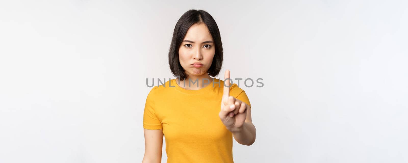 Portrait of asian woman looking serious and angry, showing stop prohibit gesture, taboo sign, forbidding smth, standing in yellow tshirt over white background.