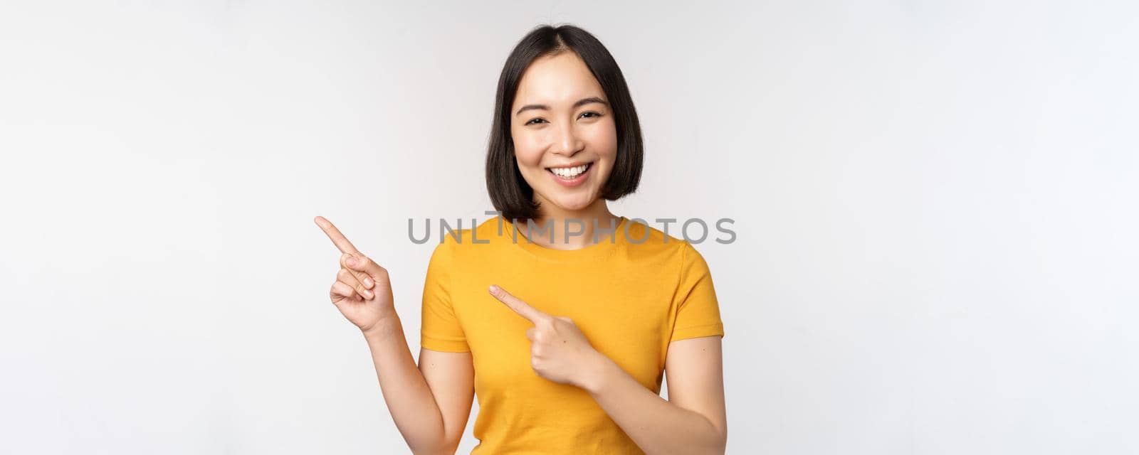 Portrait of smiling asian brunette girl in yellow tshirt, pointing fingers left, showing copy space, promo deal, demonstrating banner, standing over white background by Benzoix