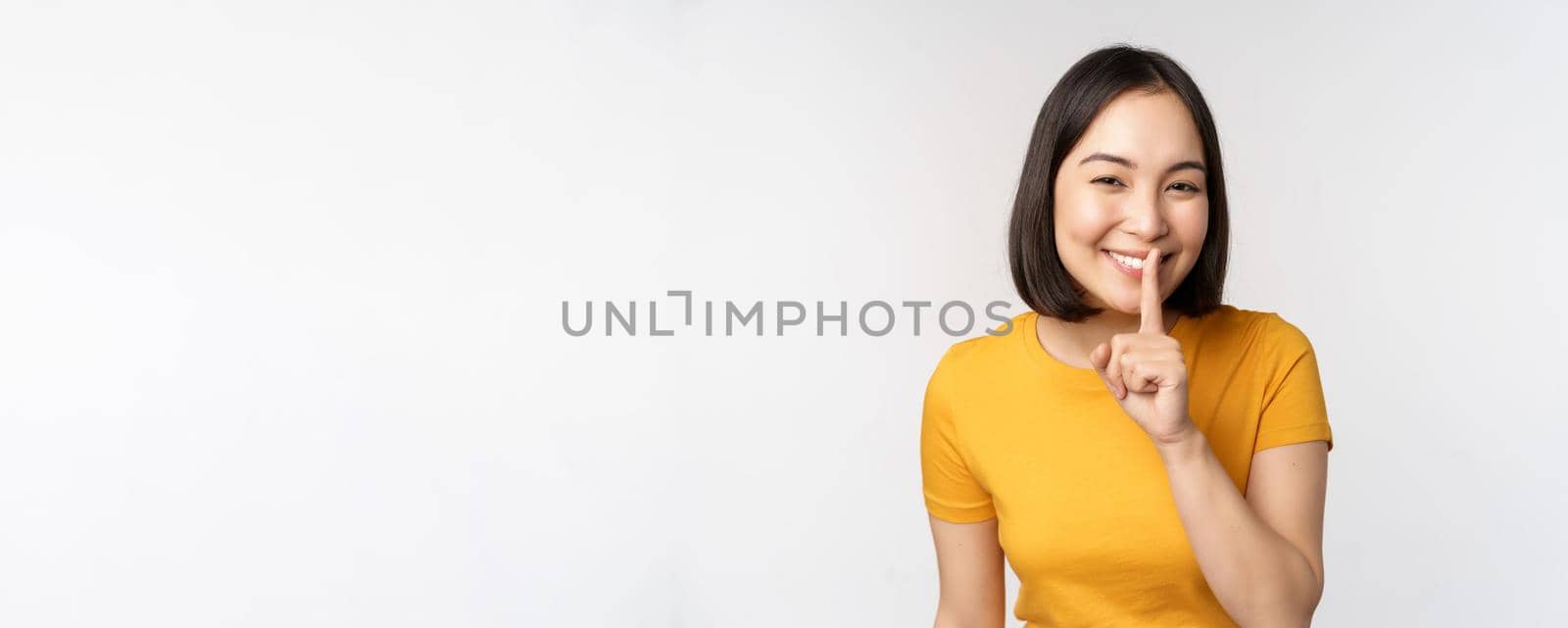 Keep quiet. Cute asian woman make shhh gest, showing shush, hush sign, press finger to lips, silence, standing over white background.
