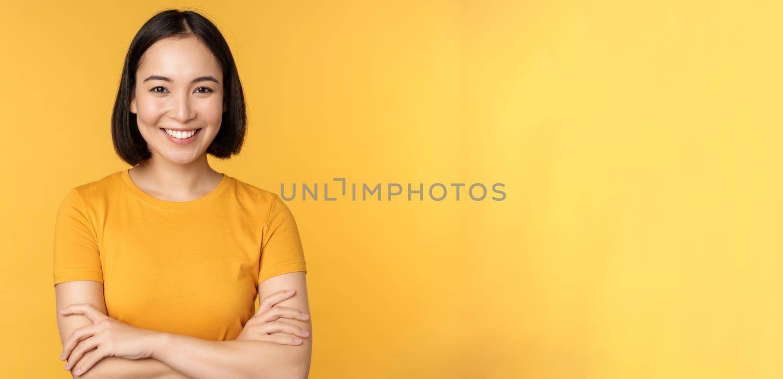 Confident asian girl cross arms on chest, smiling and looking assertive, standing over yellow background by Benzoix