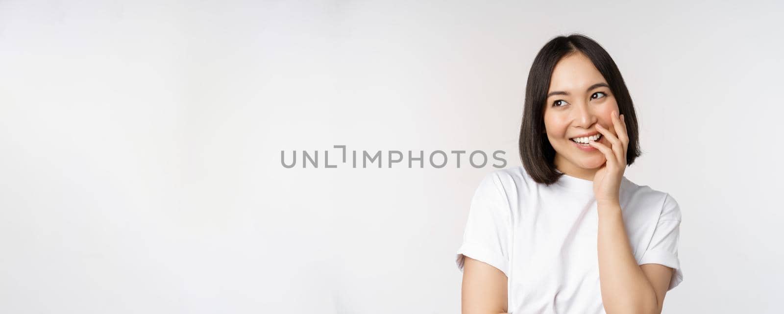 Portrait of young beautiful woman, korean girl laughing and smiling, looking coquettish, standing against white background.