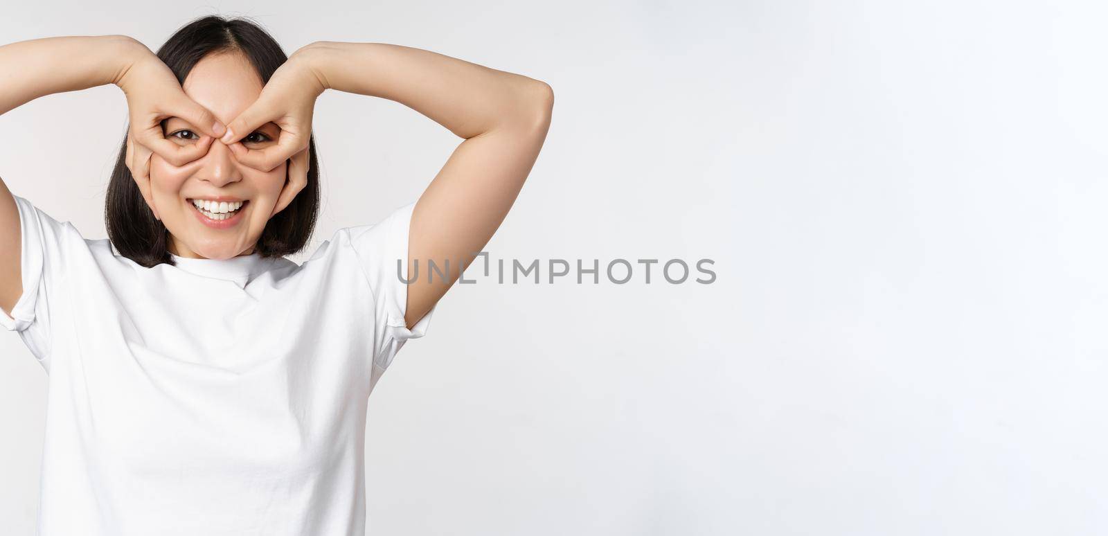 Funny young asian woman, korean girl making eyes glasses gesture, looking happy at camera, standing over white background.