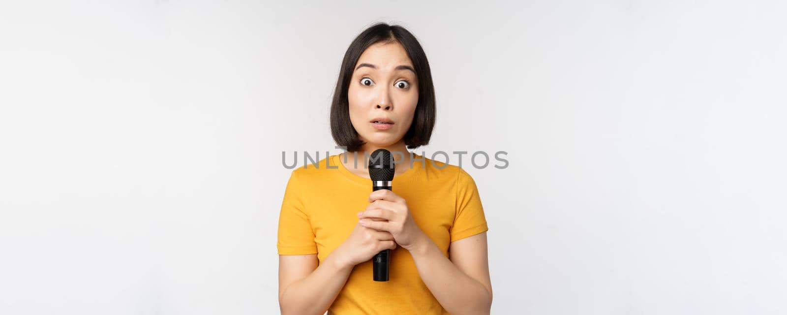 Modest asian girl holding microphone, scared talking in public, standing against white background. Copy space