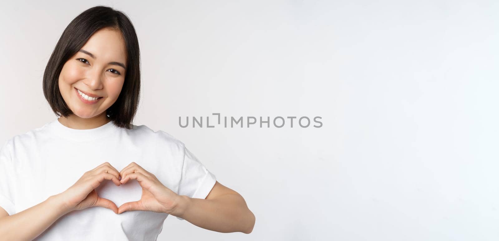 Lovely asian woman smiling, showing heart sign, express tenderness and affection, standing over white background.