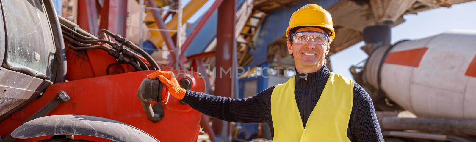 Cheerful man in work vest and safety helmet looking at camera and smiling while placing hand on truck