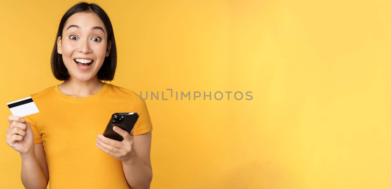 Online shopping. Cheerful asian girl holding credit card and smartphone, paying, order with mobile phone, standing over yellow background by Benzoix