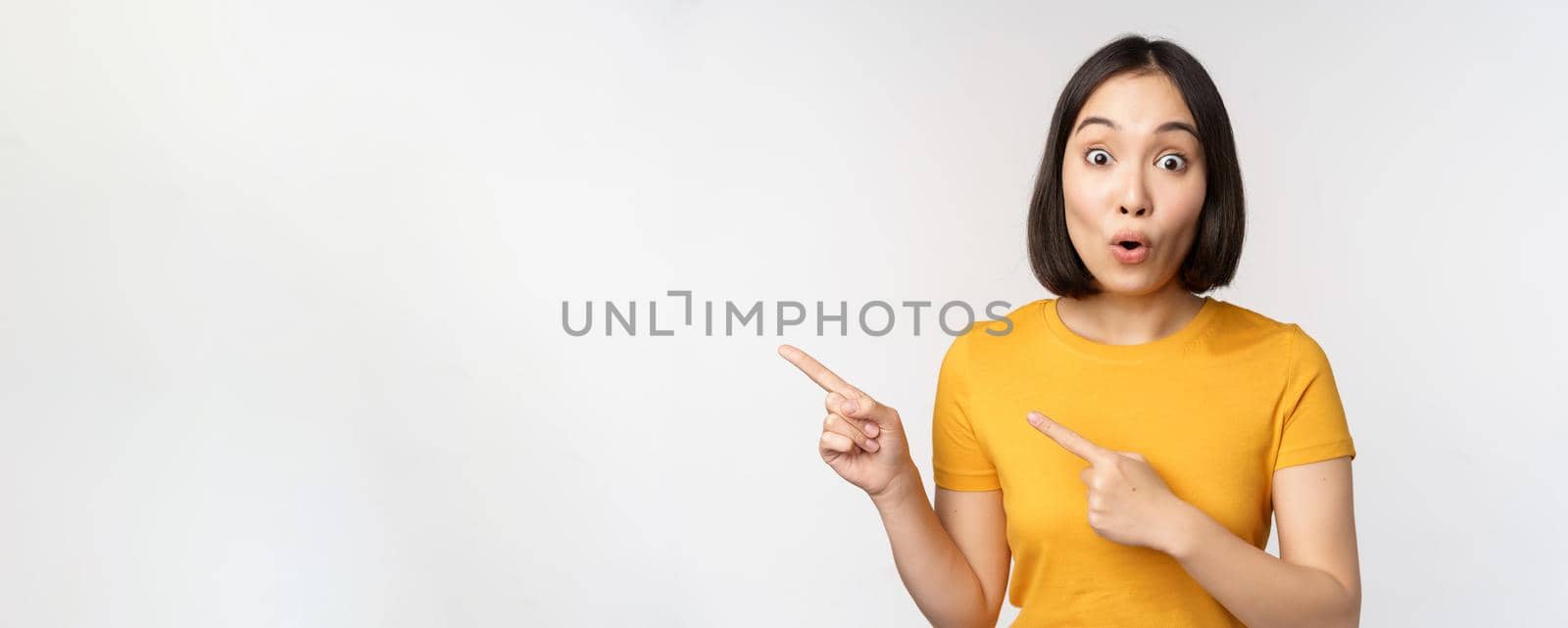 Portrait of smiling asian brunette girl in yellow tshirt, pointing fingers left, showing copy space, promo deal, demonstrating banner, standing over white background by Benzoix