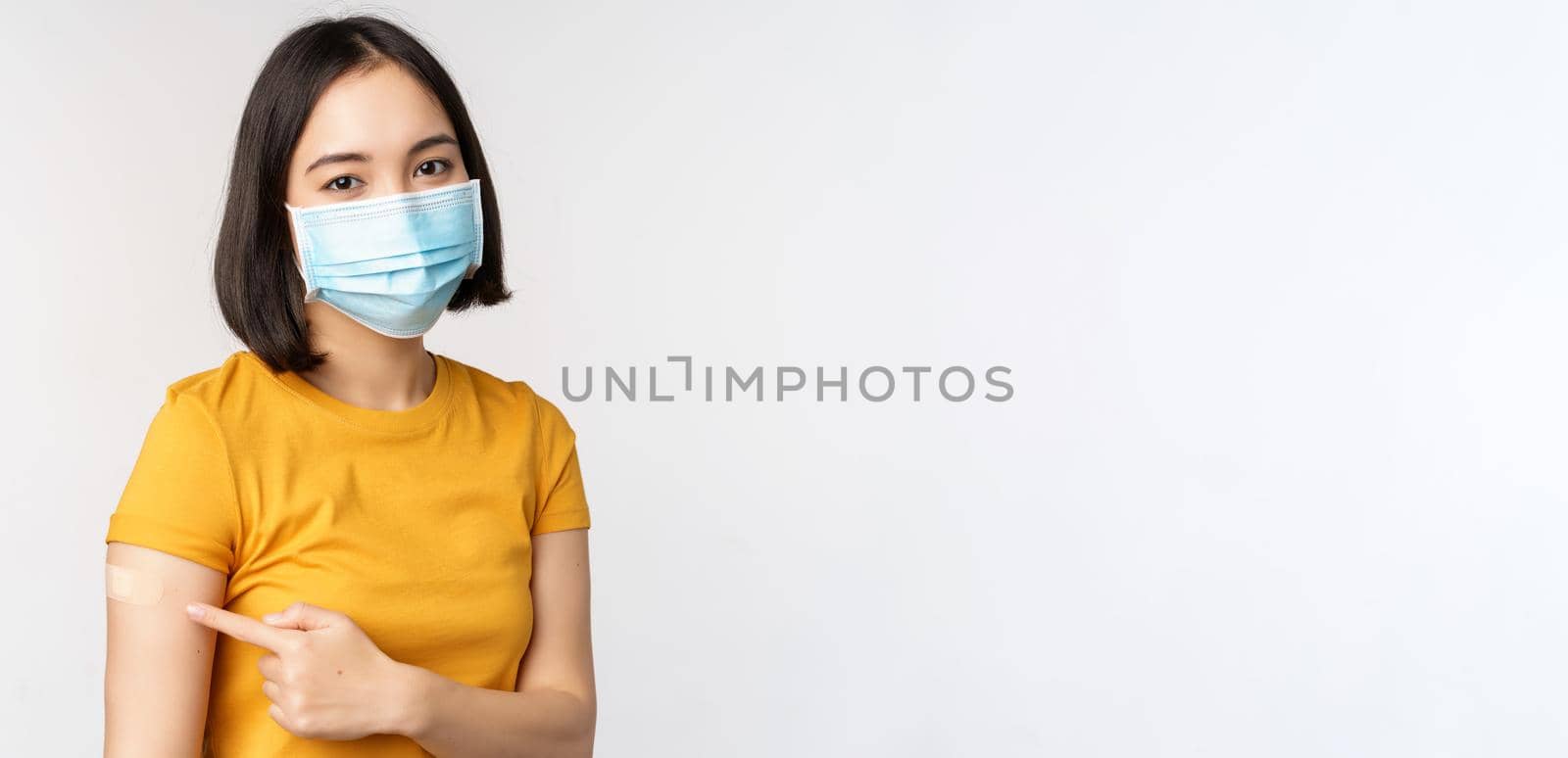 Covid-19, vaccination and healthcare concept. Portrait of cute asian girl in medical mask, has band aid on shoulder after coronavirus vaccine, standing over white background.