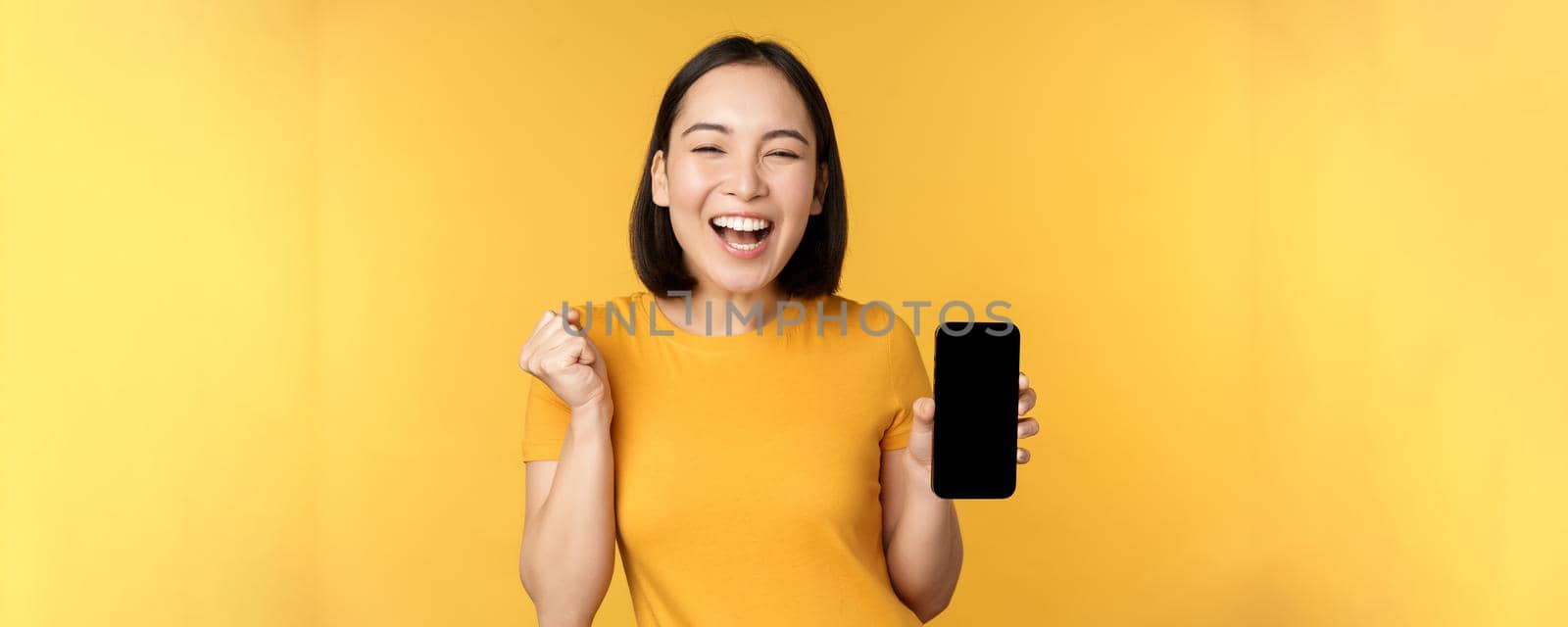 Excited asian woman, showing smartphone app and triumphing, celebrating on mobile phone, standing over yellow background. Copy space