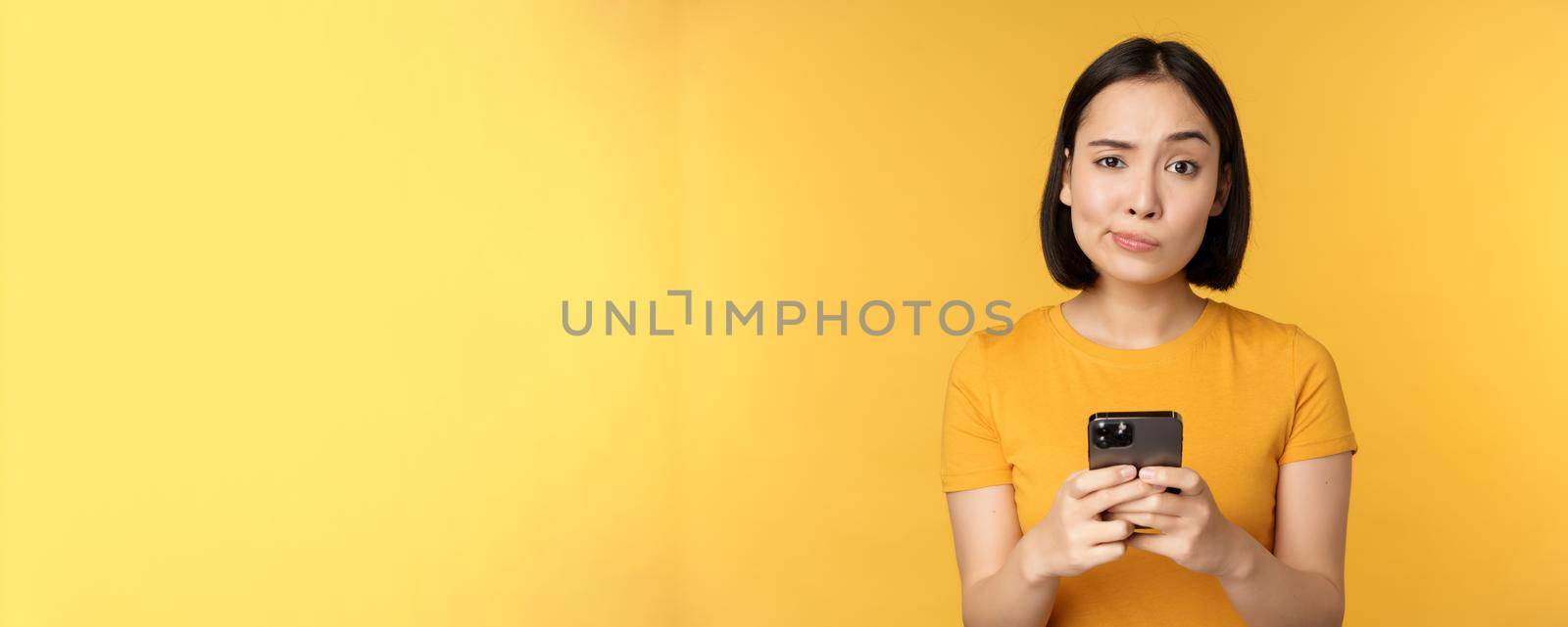 Skeptical asian woman holding smartphone, looking with doubt at camera, standing over yellow background by Benzoix