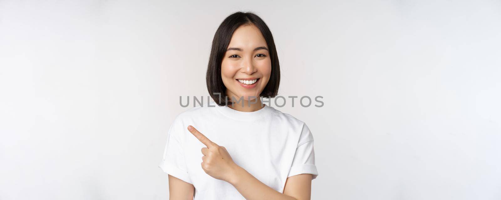 Close up of young japanese woman smiling, pointing finger left at copy space, showing announcement or advertisment banner, standing over white background by Benzoix