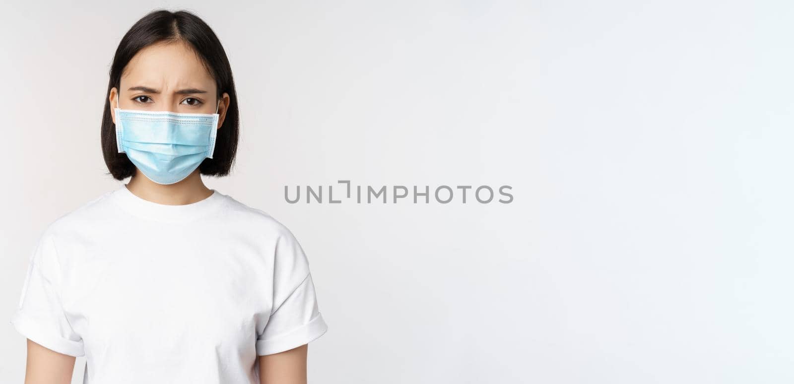 Health and covid pandemic concept. Angry young asian girl in face medical mask, frowning displeased, standing over white background.