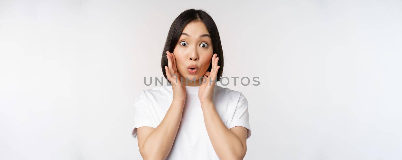 Close up portrait of asian girl looking surprised, wow face, reacting amazed at smth, standing in white tshirt over studio background, isolated.