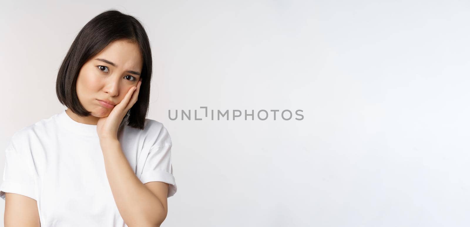 Sad asian girl looking upset and lonely, sulking and frowning, standing against white background in casual tshirt by Benzoix