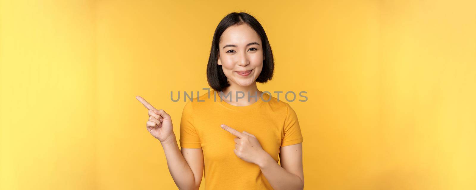 Smiling asian woman pointing fingers left, showing advertisement on empty copy space, standing over yellow background by Benzoix