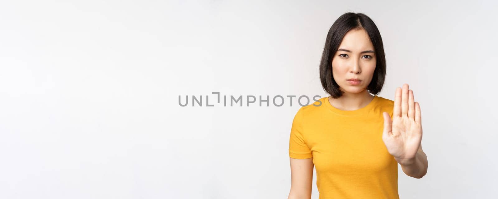 Portrait of asian woman looking serious and angry, showing stop prohibit gesture, taboo sign, forbidding smth, standing in yellow tshirt over white background by Benzoix