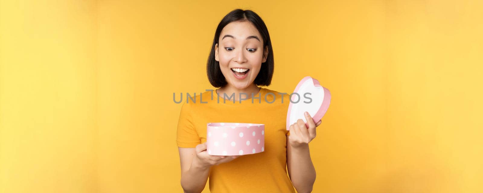 Portrait of excited asian woman, open gift box with surprised happy face, standing over yellow background.