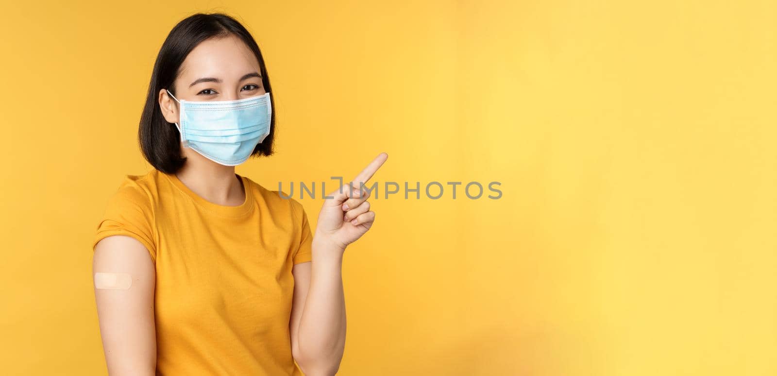 Vaccination from covid and health concept. Image of smiling korean girl in medical face mask, band aid on shoulder, pointing finger at banner advertisement, yellow background by Benzoix