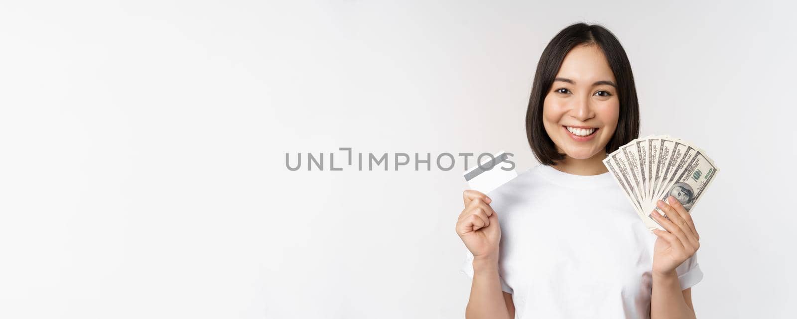 Portrait of asian woman smiling, holding credit card and money cash, dollars, standing in tshirt over white background by Benzoix