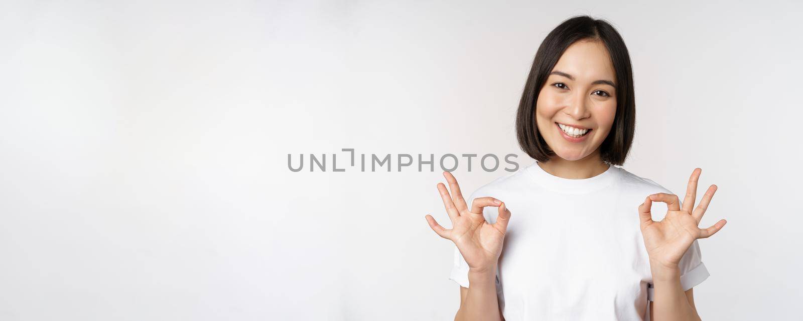 Very well, excellent. Smiling asian woman showing okay sign, approval, ok gesture, looking satisfied, recommending smth, standing over white background by Benzoix