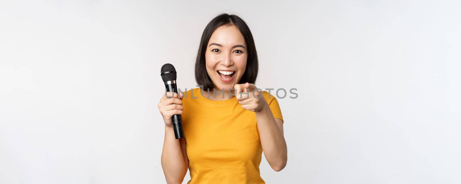 Enthusiastic asian girl with microphone, pointing finger at camera, suggesting you to sing, standing over white background.