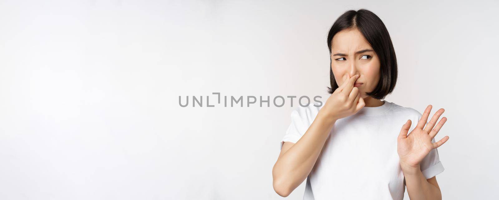 Asian girl looks disgusted, rejecting product with bad smell, shut nose from aversion and cringe, standing against white background.