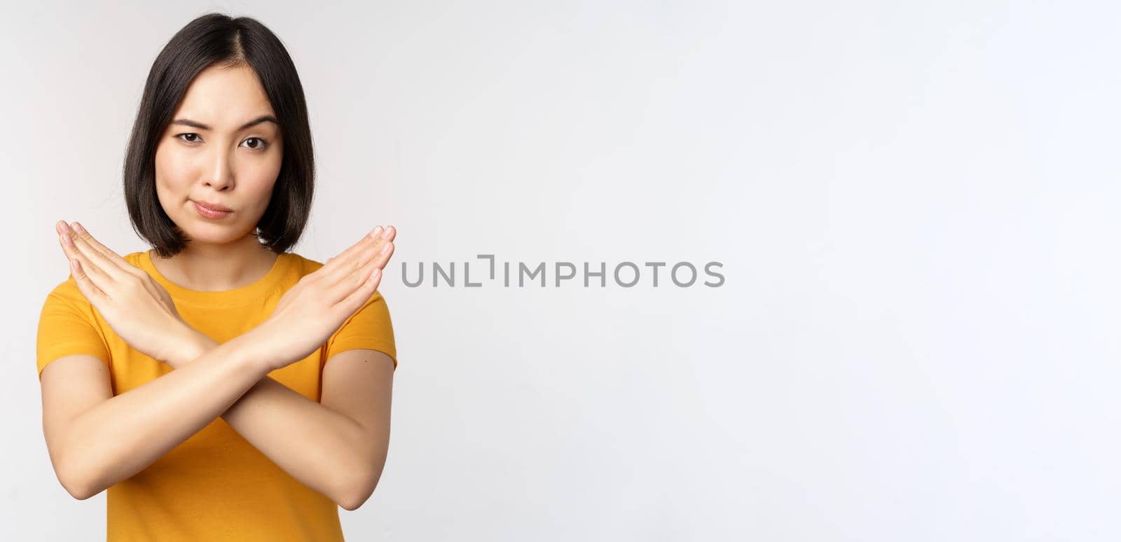 Portrait of asian woman looking serious and angry, showing stop prohibit gesture, taboo sign, forbidding smth, standing in yellow tshirt over white background by Benzoix