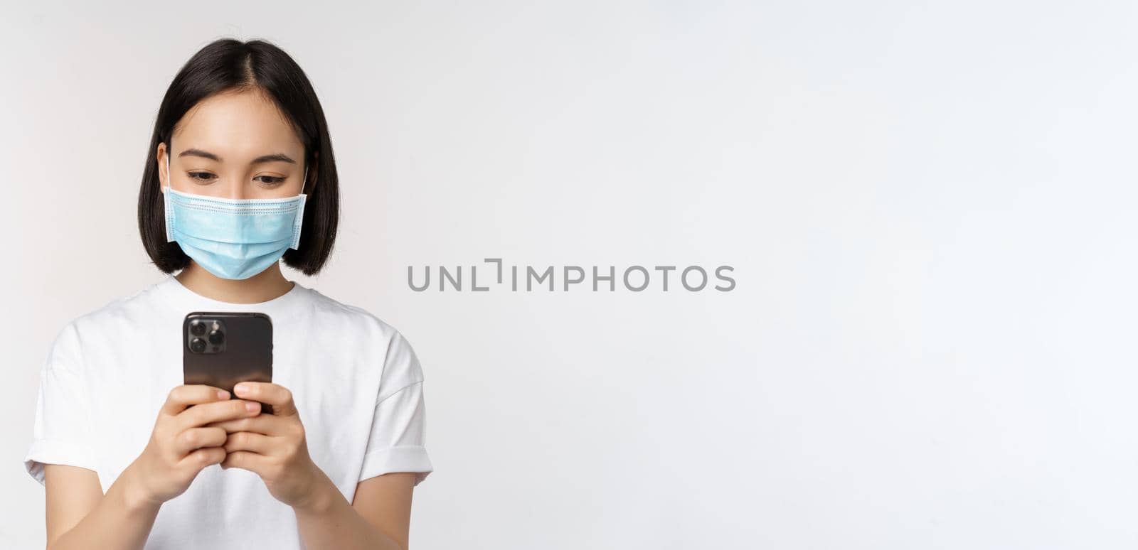 Health, covid and mobile concept. Young asian woman in medical face mask, looking at smartphone screen, using phone app, shopping online, white background.