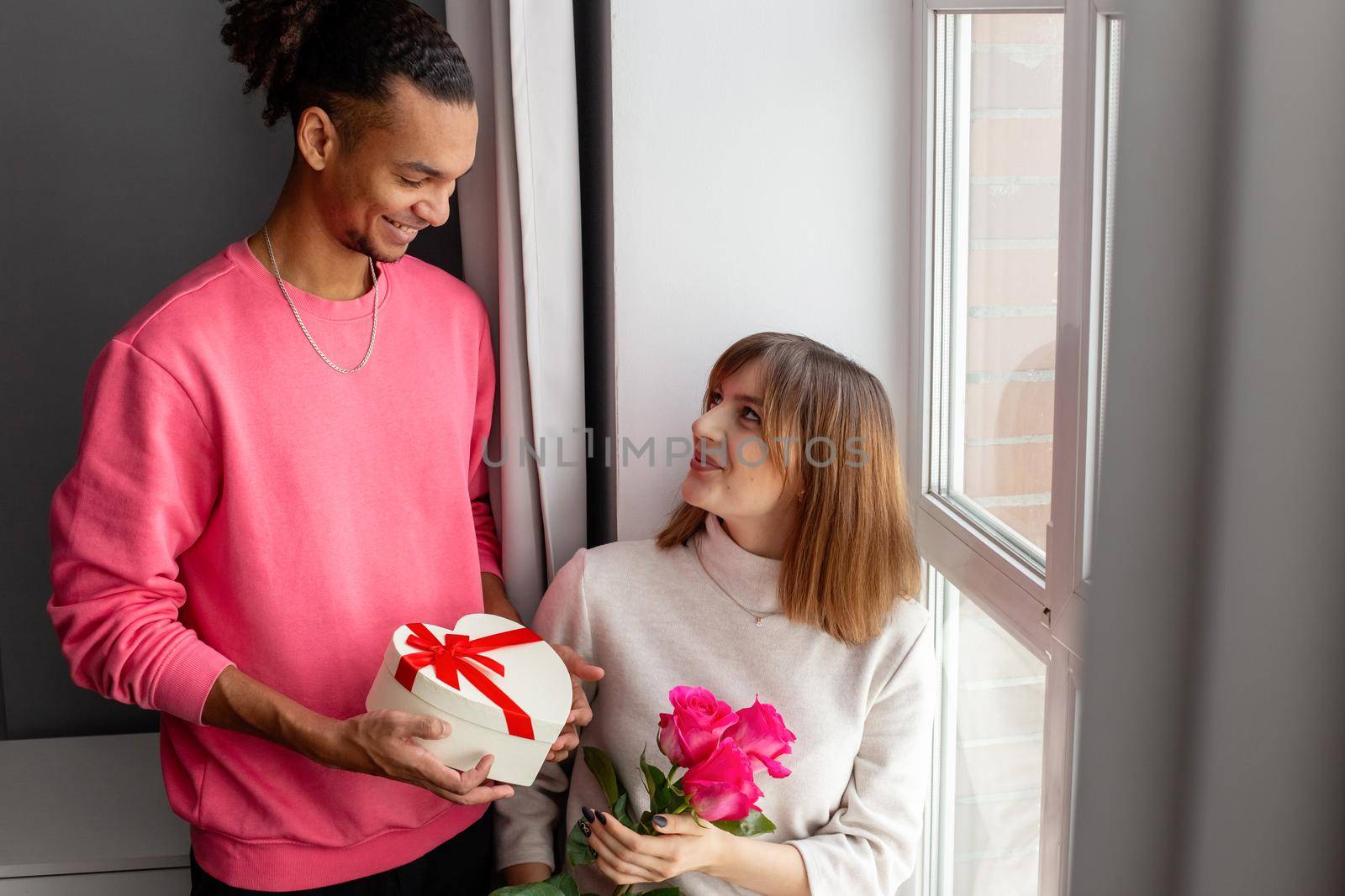 A guy in a pink jumper gives his girlfriend, sitting by the window, a white heart-shaped box with a bow and a bouquet of roses. Copy space