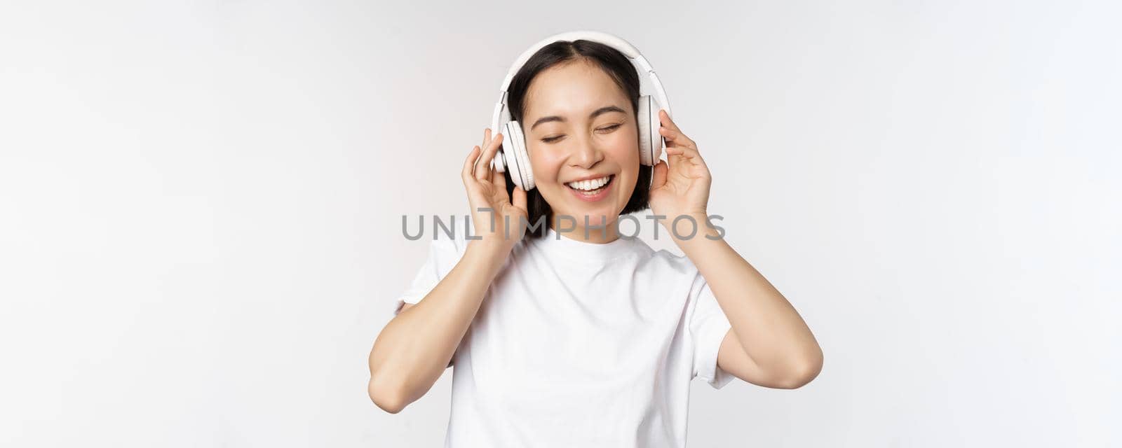 Modern asian girl dancing, listening music with headphones, smiling happy, standing in tshirt over white background.