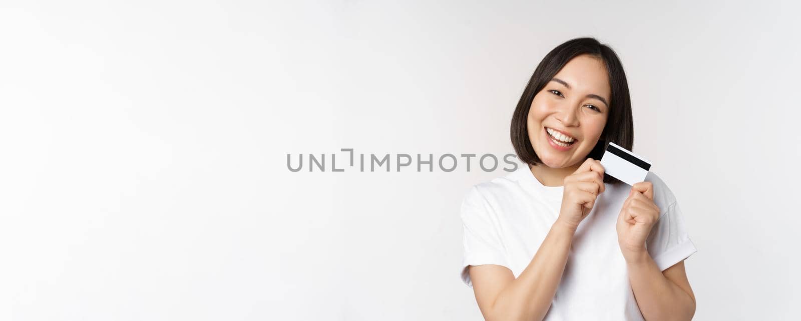 Image of smiling asian woman hugging credit card, buying contactless, standing in white tshirt over white background by Benzoix