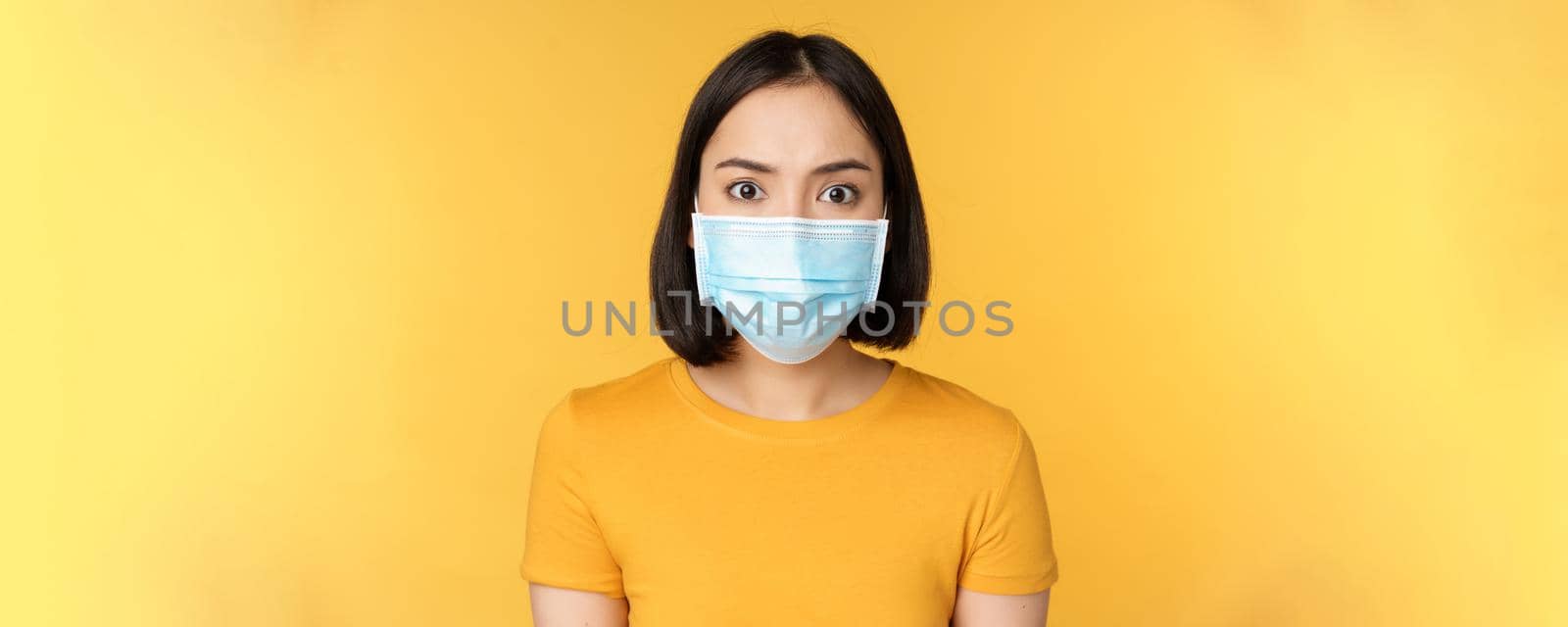 Portrait of shocked asian woman looking concerned and startled at camera, wearing covid-19 medical face mask, standing against yellow background.