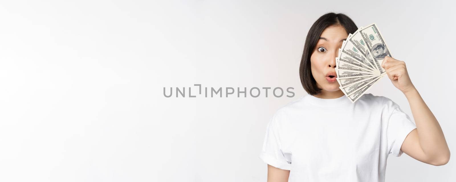 Happy asian girl holding money, cash near face, concept of microcredit and finance, white studio background by Benzoix