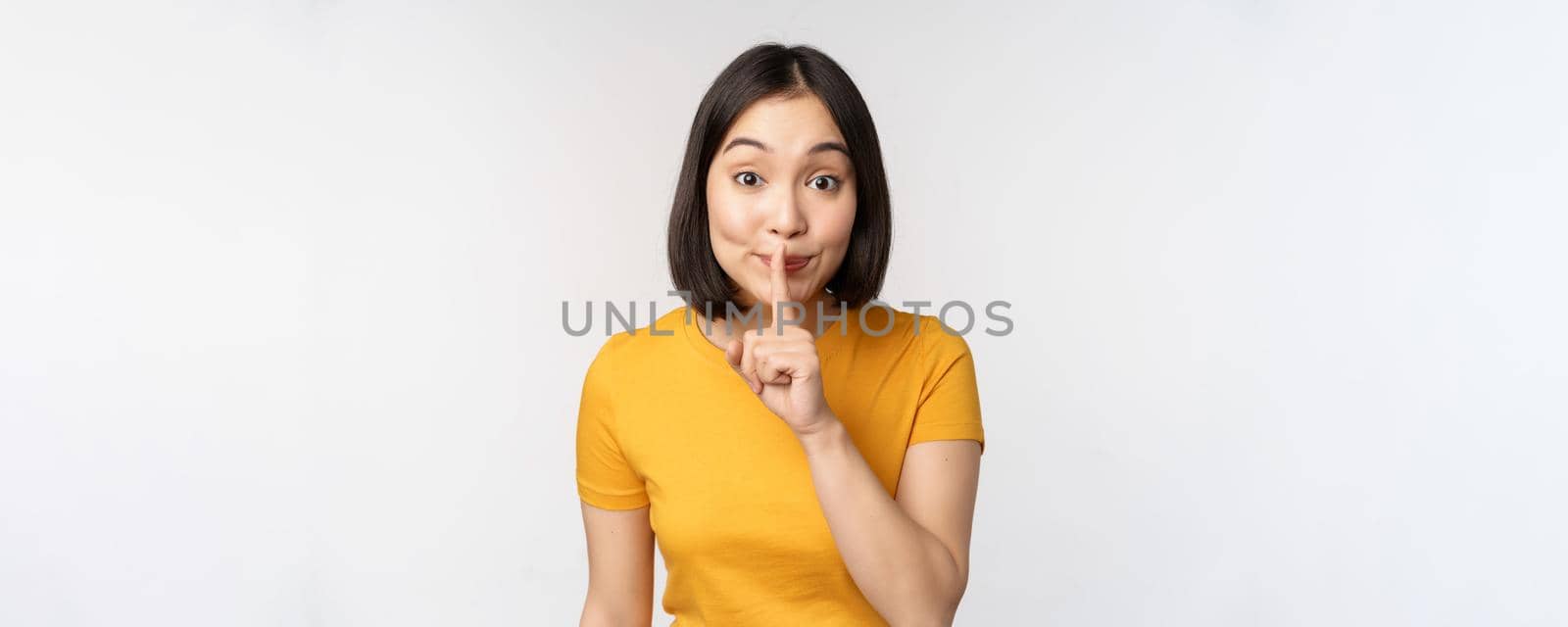 Keep quiet. Cute asian woman make shhh gest, showing shush, hush sign, press finger to lips, silence, standing over white background.