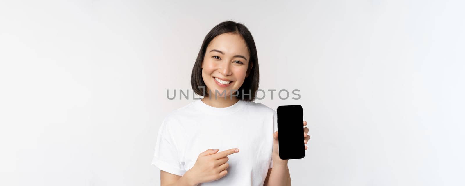 Smiling asian woman pointing finger at smartphone screen, showing application interface, mobile phone website, standing over white background.